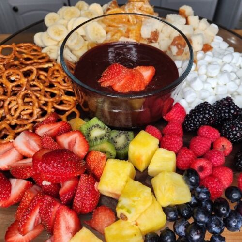 A platter of colorful fruit, pretzels, pound cake and marshmallows surrounding a bowl of chocolate fondue.