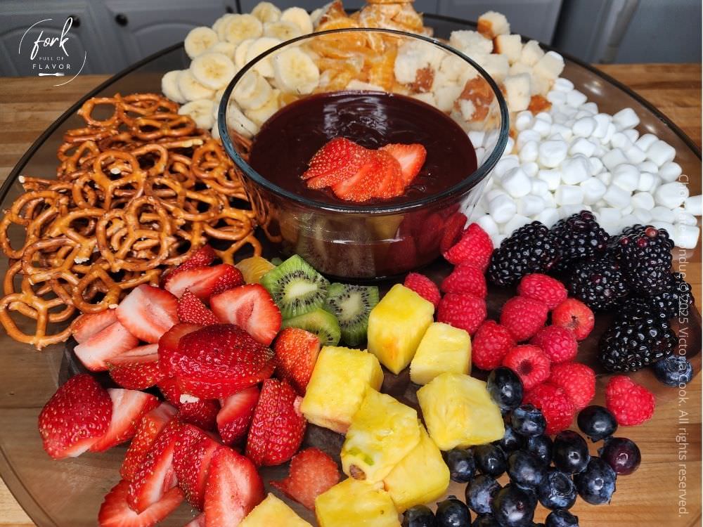 A platter of colorful fruit, pretzels, pound cake and marshmallows surrounding a bowl of chocolate fondue.