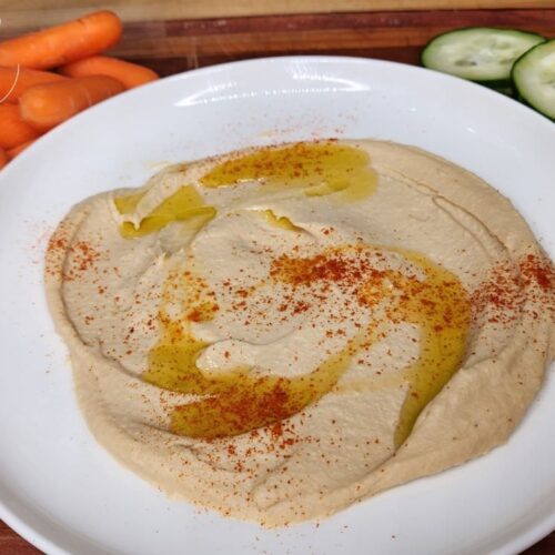 Homemade hummus plated and topped with a drizzle of olive oi l and a couple shakes of paprika ready to eat with some baby carrots and sliced cucumbers.