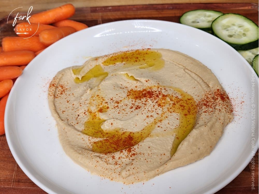 Homemade hummus plated and topped with a drizzle of olive oi l and a couple shakes of paprika ready to eat with some baby carrots and sliced cucumbers.